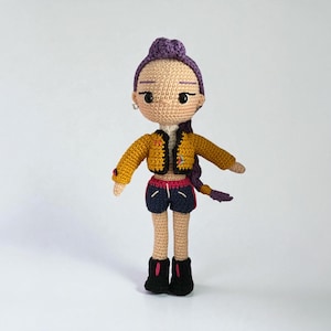 May include: Crocheted doll with purple hair styled in a bun and braid. The doll wears a mustard yellow jacket, navy blue shorts, and black boots. The jacket has a star patch. The doll has a neutral skin tone and is wearing silver hoop earrings.
