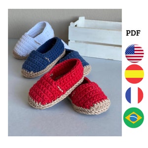 Puede incluir: Tres pares de zapatos de bebé tejidos a crochet en blanco, azul y rojo. Los zapatos tienen una suela beige y están colocados sobre una superficie de madera.