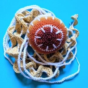 May include: A handmade crocheted ball, in shades of orange and brown with white embroidered details, rests on a crocheted net. The net has a spiral design in tan and white. The background is bright blue. The item is likely a decorative piece.