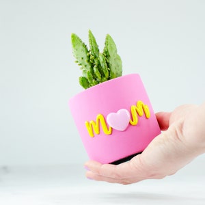 Könnte beinhalten: Ein rosa Blumentopf mit dem Wort "MOM" in gelben Buchstaben und einem rosa Herzen in der Mitte. Eine kleine grüne Kaktuspflanze wächst im Topf.