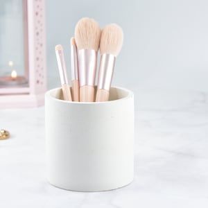 May include: A white concrete makeup brush holder with five brushes inside. The brushes have pink and gold handles and soft, white bristles.
