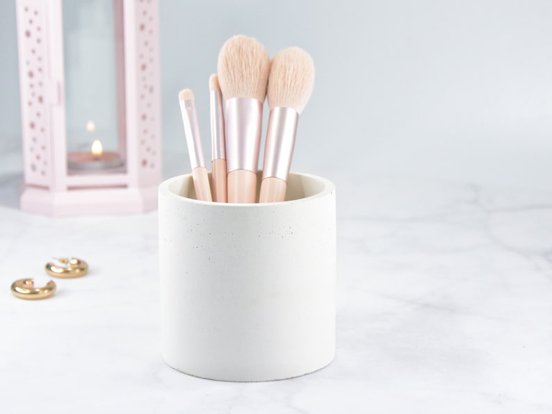 May include: A white concrete makeup brush holder with five brushes inside. The brushes have pink and gold handles and soft, white bristles.