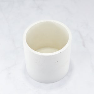 May include: A white cylindrical concrete planter with a smooth, slightly textured surface. The planter is empty and ready for a plant.