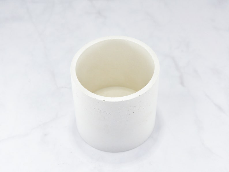 May include: A white cylindrical concrete planter with a smooth, slightly textured surface. The planter is empty and ready for a plant.
