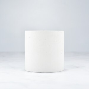 May include: A white concrete planter with a speckled finish. The planter is cylindrical in shape and has a smooth, rounded edge.