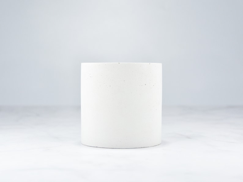 May include: A white concrete planter with a speckled finish. The planter is cylindrical in shape and has a smooth, rounded edge.