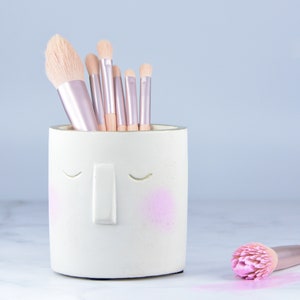 Könnte beinhalten: Ein weißer Keramik-Make-up-Pinselhalter in Form eines Gesichts mit geschlossenen Augen und einer Nase. Der Halter hat rosafarbene Rouge-Flecken auf den Wangen. Sieben Make-up-Pinsel befinden sich im Halter. Ein rosafarbener Make-up-Pinsel liegt auf einer weißen Marmorfläche.