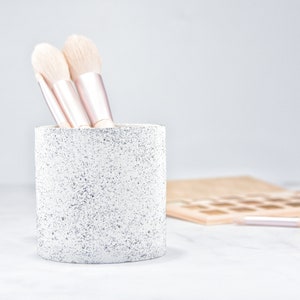 Könnte beinhalten: Ein weißer Keramik-Make-up-Pinselhalter mit gesprenkeltem Finish. Drei rosafarbene und weiße Make-up-Pinsel befinden sich im Halter.