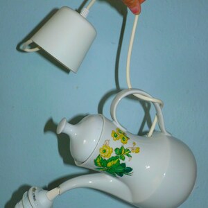 Könnte beinhalten: Eine weiße Porzellan-Teekanne mit Blumenmuster wird als Hängelampe verwendet. Die Teekanne hat ein grünes und gelbes Blumenmuster. Die Leuchte hängt an einem weißen Kabel.