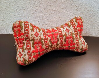 May include: Bone-shaped cushion with a red, green, and cream floral pattern. The cushion is made of a textured fabric and sits on a dark brown surface. The design features vertical stripes with floral motifs.