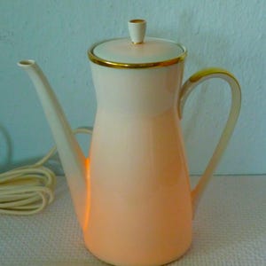 Puede incluir: Una cafetera eléctrica de cerámica blanca y melocotón con un borde dorado alrededor de la tapa y el asa. La cafetera tiene un cable y está iluminada desde el interior.