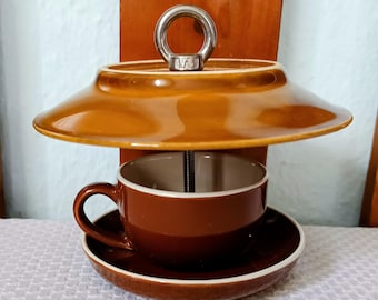 Op de afbeelding: Een bruine keramische vogelvoeder met een kop-en-schotel ontwerp. De voeder heeft een brede, schotelvormige bovenkant en een kopje dat eronder hangt. Een metalen ring is aan de bovenkant bevestigd om op te hangen. De kop en schotel zijn bruin met een witte rand.
