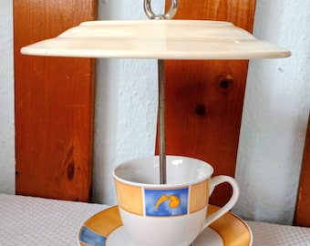 Op de afbeelding: Een decoratieve kop en schotel set met een crèmekleurig, parapluvormig deksel en een metalen staaf. De kop en schotel hebben oranje en blauwe accenten. De kop heeft een blauw vierkant met een geel ontwerp.