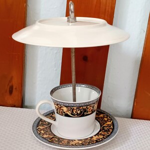 Könnte beinhalten: Ein dekoratives Tassen- und Untertassen-Set mit einem passenden Teller darüber. Die Tasse, die Untertasse und der Teller haben einen weißen Hintergrund mit schwarzen und goldenen Zierdesigns. Eine Metallstange verbindet die Tasse mit dem Teller.