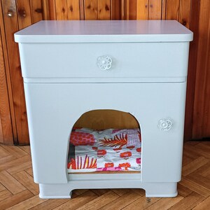 Può includere: Un comodino in legno dipinto di azzurro con un cassetto e un'apertura per un letto per animali domestici. Il cassetto ha una manopola a forma di fiore bianco. L'apertura contiene un cuscino con motivi colorati.