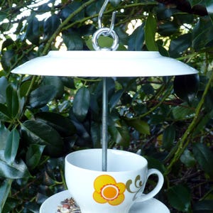 Könnte beinhalten: Eine weiße Teetasse aus Keramik mit einem gelben Blumendesign wird als Vogel futterspender verwendet. Die Teetasse ist an einer weißen Untertasse und einem weißen Teller mit einem Metallhaken befestigt. Der Futterspender hängt an einem Baumzweig.