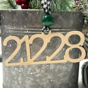 Puede incluir: Un adorno de madera con el año 2023 recortado en una fuente decorativa. El adorno cuelga de una cinta a cuadros blanco y negro con una cuenta verde.