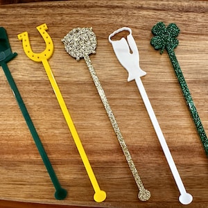 May include: Five St. Patrick's Day themed drink stirrers. The stirrers include a green top hat, yellow horseshoe, gold glitter ball, white beer mug, and green glitter shamrock. The stirrers are on a wooden surface.