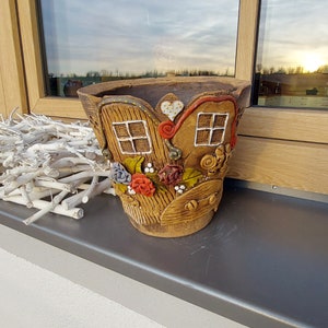 Könnte beinhalten: Ein brauner Keramik-Pflanztopf mit einem skulpturalen Hausdesign, das zwei Fenster, ein Herz und eine Tür zeigt. Die Außenseite ist mit bunten Tonblumen in Rot, Blau und Grün sowie mit Holzmaserungsmustern und kleinen weißen Punkten verziert.