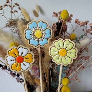 Könnte beinhalten: Drei blumenförmige Holzdekorationen auf dünnen Stäben. Eine Blume ist hellblau mit gelber Mitte, eine ist gelb-weiß mit orangefarbener Mitte und eine ist hellgrün mit gelber Mitte. Jedes Teil hat eine glatte, glänzende Oberfläche.