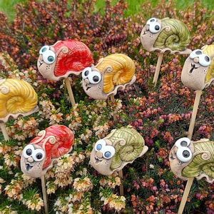 Könnte beinhalten: Ein Set aus drei Keramikbechern mit einer gesprenkelten, cremefarbenen Glasur. Jeder Becher hat ein anderes handgemaltes Design: ein blaues und grünes Blatt, eine gelbe Blume und eine rote Beere. Die Becher haben ein schlichtes, modernes Design.