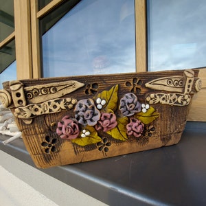 Könnte beinhalten: Ein rechteckiger Keramik-Blumentopf mit rustikalem, verwittertem Aussehen. Er weist geschnitzte Blumen- und geometrische Muster sowie erhabene Rosen und Blätter auf. Die Farbpalette umfasst Braun-, Lila-, Rosa- und Grüntöne.