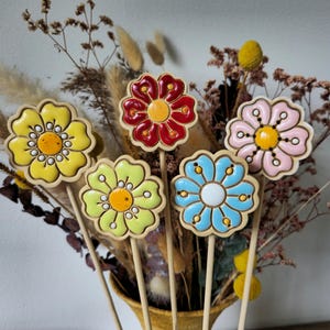 Könnte beinhalten: Eine Sammlung farbenfroher Gartenstecker aus Keramik in Blumenform. Die Stecker zeigen Blumendesigns in Gelb, Rot, Rosa, Blau und Grün mit gelben und weißen Akzenten. Die Stecker sind auf Holzstäben und in einer Vase mit Trockenblumen angeordnet.