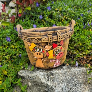 Könnte beinhalten: Dekorativer, handbemalter Keramik-Pflanztopf mit korbähnlichem Design. Der Pflanztopf zeigt zwei Cartoon-Kuhfiguren mit gelben und roten Akzenten. Der Pflanztopf hat zwei Griffe.