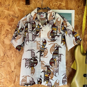 May include: A short-sleeved button-down shirt in white featuring a repeating graphic print of stylised sailboats in shades of brown, yellow, and black. The shirt has a classic collar, a front button placket, and a single chest pocket on the left side.