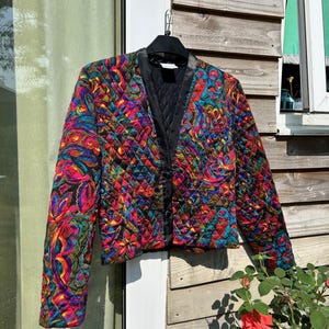 May include: A quilted jacket featuring a vibrant, multi-coloured paisley and floral pattern in shades of hot pink, electric blue, yellow, and black. The jacket has a V-neckline, long sleeves, and a solid black satin-like trim along the front opening.