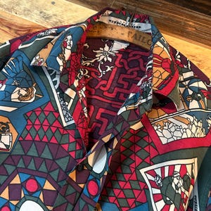 May include: A red, blue and green patterned button-up shirt with a geometric design. The shirt has a collar and a button closure. The fabric is a lightweight cotton blend.