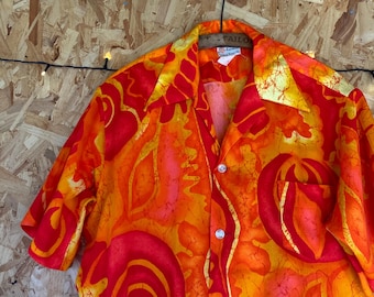 May include: A short-sleeved button-down shirt with a vibrant orange and red abstract print. The shirt has a collar and a pocket on the left side. The label on the shirt reads "Tailored in Hawaii".