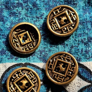 May include: Three round, metallic gold-toned buttons featuring a raised square center with two holes for attachment. The surface of each button is decorated with embossed geometric patterns and symbols surrounding the central square frame.