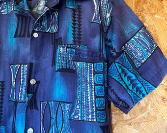 May include: A short-sleeved button-down shirt in shades of blue with a geometric pattern. The shirt features a pocket and white buttons. The design includes shapes and lines in teal and white against a darker blue background.