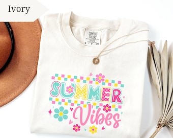 May include: Ivory-colored t-shirt with the words "SUMMER VIBES" in a colorful, floral design. The shirt has a crew neck and short sleeves. A gold necklace with a pendant is draped across the shirt.