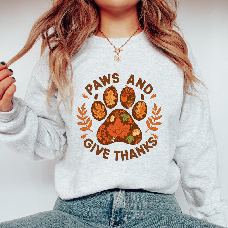 Peut inclure: Un sweat-shirt gris clair avec un motif de patte de chien rempli de feuilles d'automne et les mots "PAWS AND GIVE THANKS" en marron. Le motif comprend des feuilles et des brins orange et marron.
