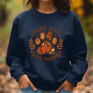 Peut inclure: Sweat-shirt bleu marine avec un motif de patte sur le th&egrave;me de l'automne et le texte "PAWS AND GIVE THANKS" en marron. L'empreinte de patte est compos&eacute;e de feuilles d'automne et d'une citrouille. Le motif est entour&eacute; d'accents de feuilles et de branches.