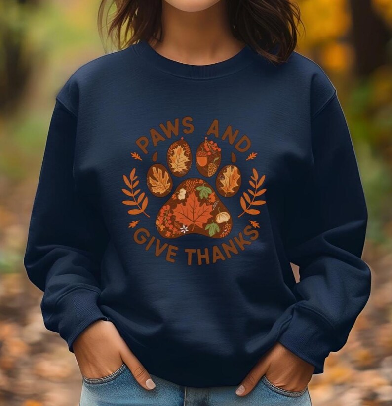 Peut inclure: Sweat-shirt bleu marine avec un motif de patte sur le th&egrave;me de l'automne et le texte "PAWS AND GIVE THANKS" en marron. L'empreinte de patte est compos&eacute;e de feuilles d'automne et d'une citrouille. Le motif est entour&eacute; d'accents de feuilles et de branches.