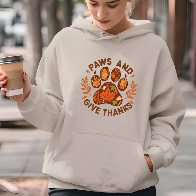 Peut inclure: Sweat &agrave; capuche cr&egrave;me avec un motif de patte rempli de feuilles d'automne et le texte "PAWS AND GIVE THANKS" en marron. Le motif comprend des feuilles orange et marron et est entour&eacute; de brins de feuilles. Le sweat &agrave; capuche a une poche avant et une capuche &agrave; cordon.