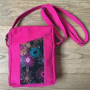 May include: A pink crossbody bag with a black and colourful patterned pocket. The bag has a polka dot pattern and a long adjustable strap.