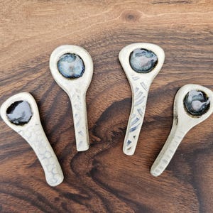May include: Four handmade ceramic spoons with a cream-colored finish and dark blue and gray accents. The spoons have a unique design with textured handles and a small, rounded bowl. They are arranged on a dark wood surface.