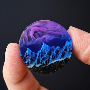 May include: A round resin pendant featuring a blue and purple mountain scene. The mountains are detailed and the resin is clear.