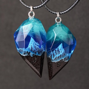 May include: Two matching pendant necklaces. Each pendant features a geometric shape with a dark brown base and a translucent blue and teal upper section, resembling a mountain range. The pendants hang from black cords.