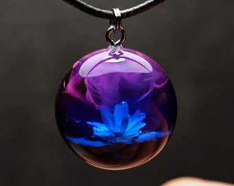 May include: A round glass pendant necklace with a black cord. The pendant contains a blue flower with pink and purple swirls in the background.