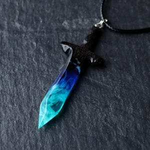 Könnte beinhalten: Ein Anhänger in Form eines kleinen Schwertes mit einem dunklen, strukturierten Griff und einer durchscheinenden Klinge, die von einem tiefen Blau am Heft zu einem hellen, wirbelnden Cyan an der Spitze übergeht, an einer schwarzen Kordel.