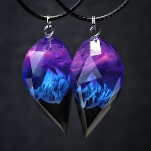 May include: Two matching pendant necklaces. Each pendant is a faceted, teardrop-shaped resin with a blue and purple landscape design. The bottom portion is black. The pendants hang from black cords with silver-coloured clasps.