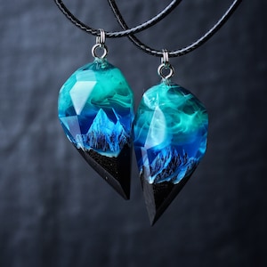 May include: Two matching pendant necklaces. Each pendant features a geometric shape with a black base, a mountain scene, and a translucent teal and blue resin top. The pendants hang from black cords.