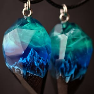 May include: Two pendants featuring a mountain scene in blue and teal resin. The faceted, gem-like pendants have a dark wood base and hang from black cords. The resin art depicts a mountain range, creating a unique jewellery piece.