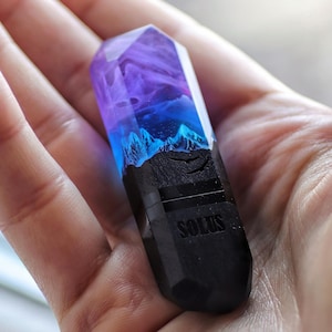 Könnte beinhalten: Ein facettiertes, kristallförmiges Objekt mit einem schwarzen Sockel, in den das Wort SOLUS eingraviert ist. Der obere Teil besteht aus durchscheinendem Harz mit einem violett-blauen Farbverlauf und einer Miniatur-Berglandschaft im Inneren.