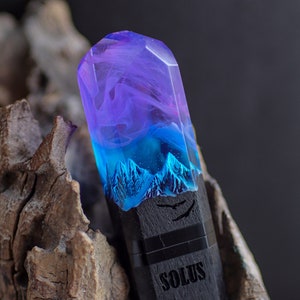 May include: A purple, blue, and black resin pendant with a mountain scene and the word "SOLUS" engraved on the bottom.